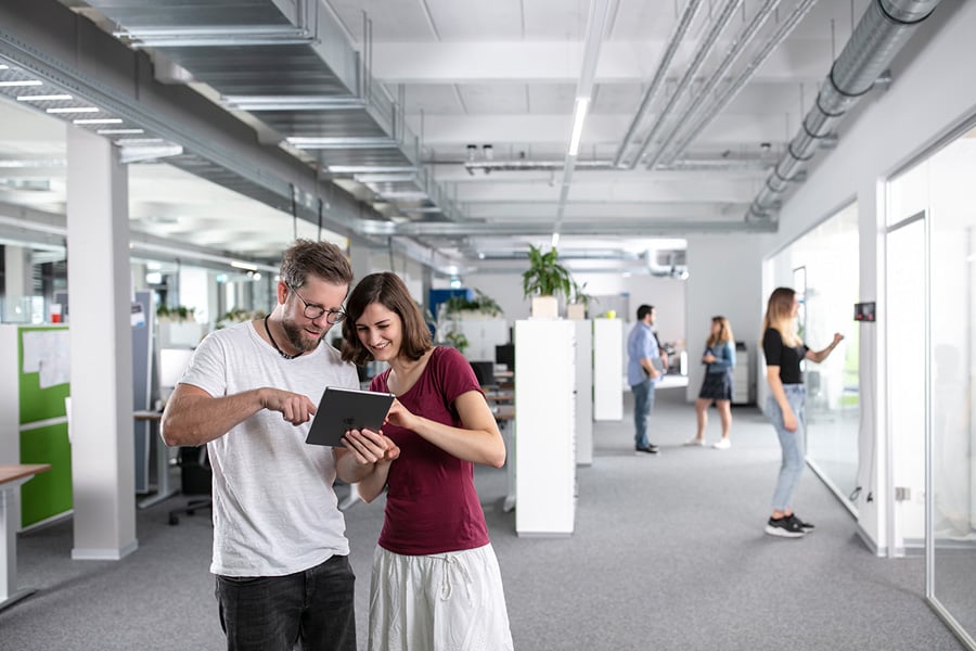 Szene im Großraumbüro. Im Vorderund stehen zwei Personen, die lächelnd auf ein iPad zeigen und schauen.