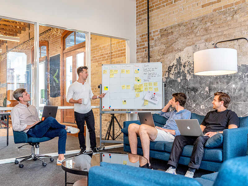 4 Männer in einem modernen Büro - 3 sitzend, 1 stehend vor dem FlipChart